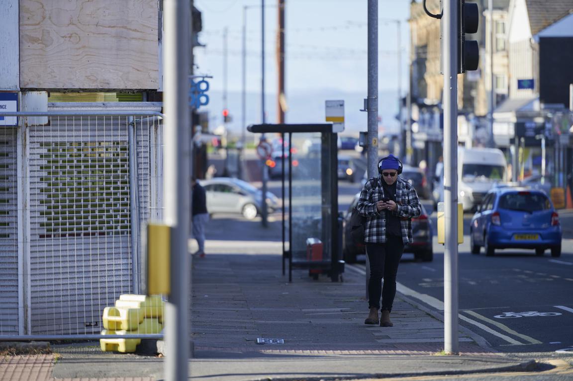 A man is walking on a sidewalk, looking down at his phone. Cars pass. The sea is in the background.