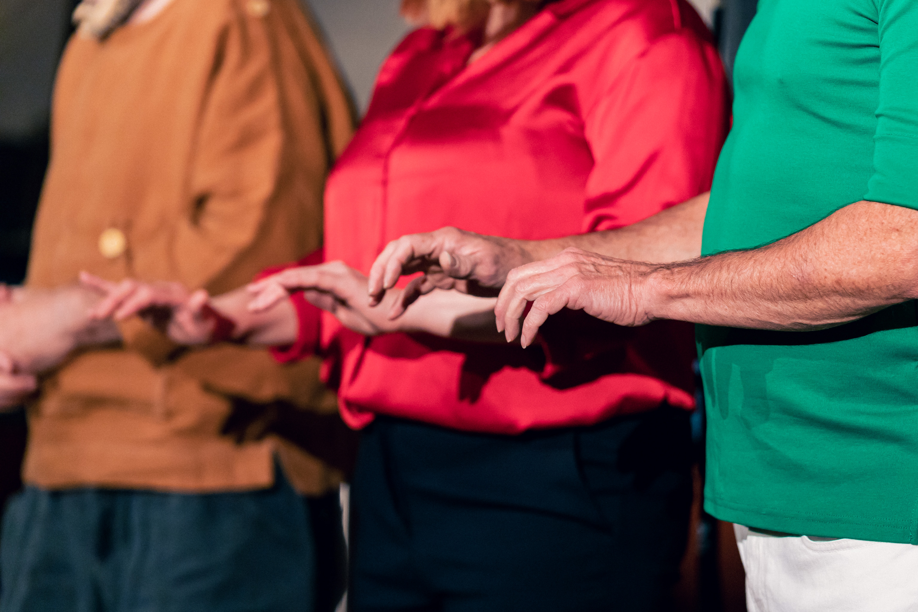 Three torsos with their hands outstretched, fingers curled and pointed downwards, as if they are all typing on keyboards. The person on the left is wearing an orange short, the person in the middle a red shirt, and the person on the right a green shirt. 