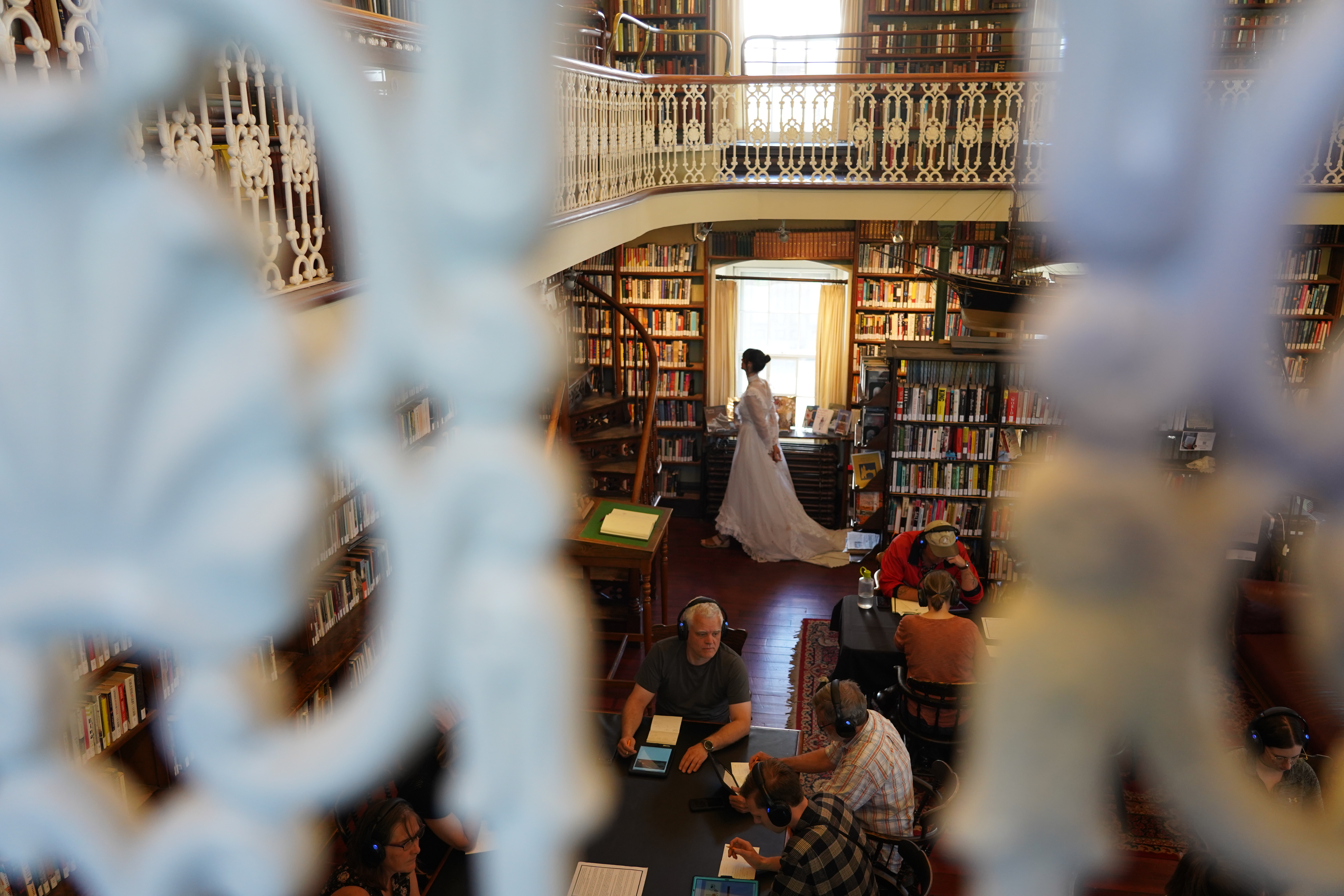 A woman in a white wedding dress walks past a window in a Victorian library. 