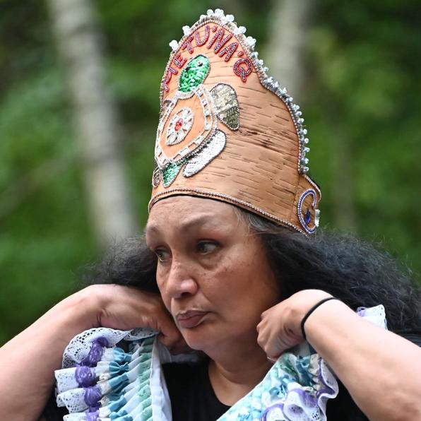 Emerald Paul, a performer in "Metu'na'q", dressed in black, adjusts a multicoloured Shakespearean Ruff around her neck, while looking intently left (out of frame). She wears a light brown Birchbark Crown which has incredible, intricate beadwork on the front. The word "Metu'na'q" (storm) at the top in red, with a turtle adorning the majority of the front of the crown. 