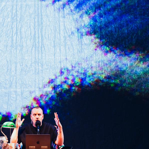 Stewart Legere, seated at a table with a computer infant of him. He hold his hands up beside his head.