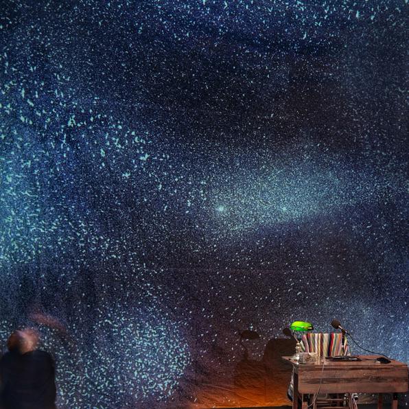 a table with a computer and green banker's light. a backdrop with projected starry sky. a man in black exits the left of the frame.