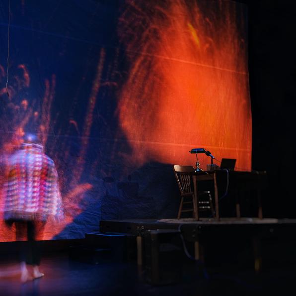 a man, wrapped in a colourful knit blanket, stands with his back to the camera. He is staring at a giant projection screen where fire and lava fill his view. Because of the angle of the shot, it sometimes appears as though he himself is inside the fire. 