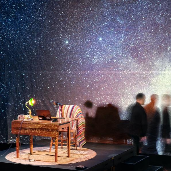 A giant projection screen with an image of the night sky, dotted with millions of stars, in blue, and purple, and black. On the left of the image, a small wooden table, with a green banker's light, a small grey rock, and an open laptop. A cozy wooden armchair is pulled up to the table, with a colourful knit blanket draped over the back. These things all sit atop a circular beige jute rug. A man, dressed in black, walks away from the table. The image is over exposed, so ghostly reflections of the man cascade out of frame.  