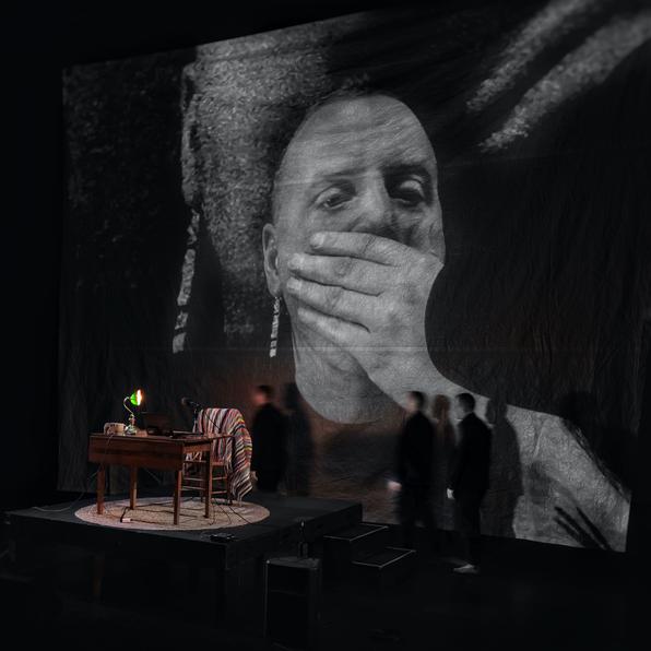 a table with a green banker's light. a chair with a rainbow blanket draped over the back. a man sits at the chair. his face is projected on the wall, very large, behind him. he is covering his mouth, as if in shock. 