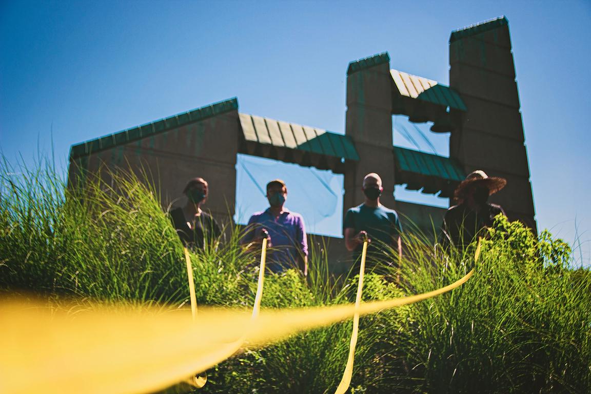 Four people in silhouette, standing in front of bell towers, holding yellow measuring tapes that extend toward to camera lens.
