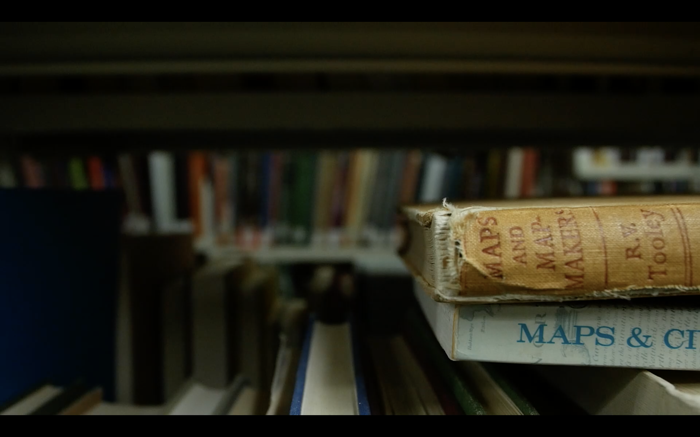Close up photograph of a library bookshelf, with blurry books in the background, and two books laying on their sides in the right foreground. The titles "Maps" and "Maps and Mapmakers" are visible on the spines of the books. 