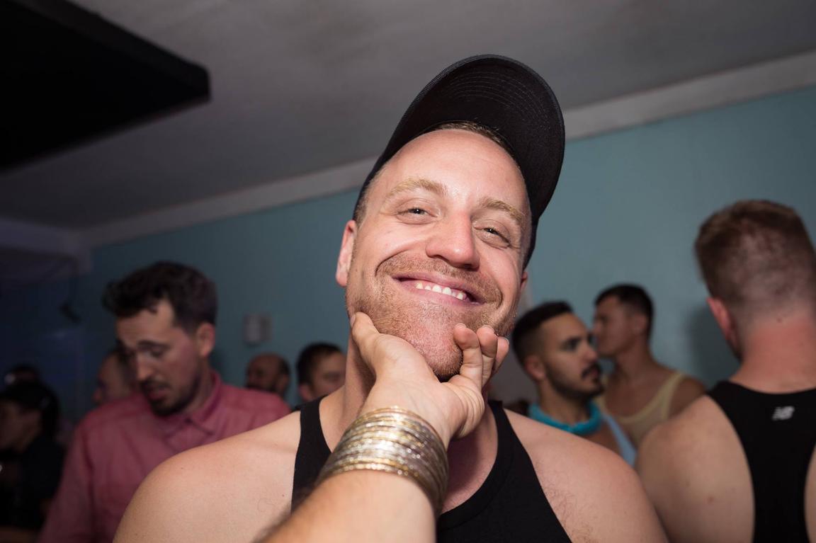A picture of Stewart Legere. An arm extends out from behind the camera and the hand is grabbing Stew's chin. Stewart is smiling and wearing a tank top and ball cap. Behind Stewart there is a group of people, maybe he is a a party?