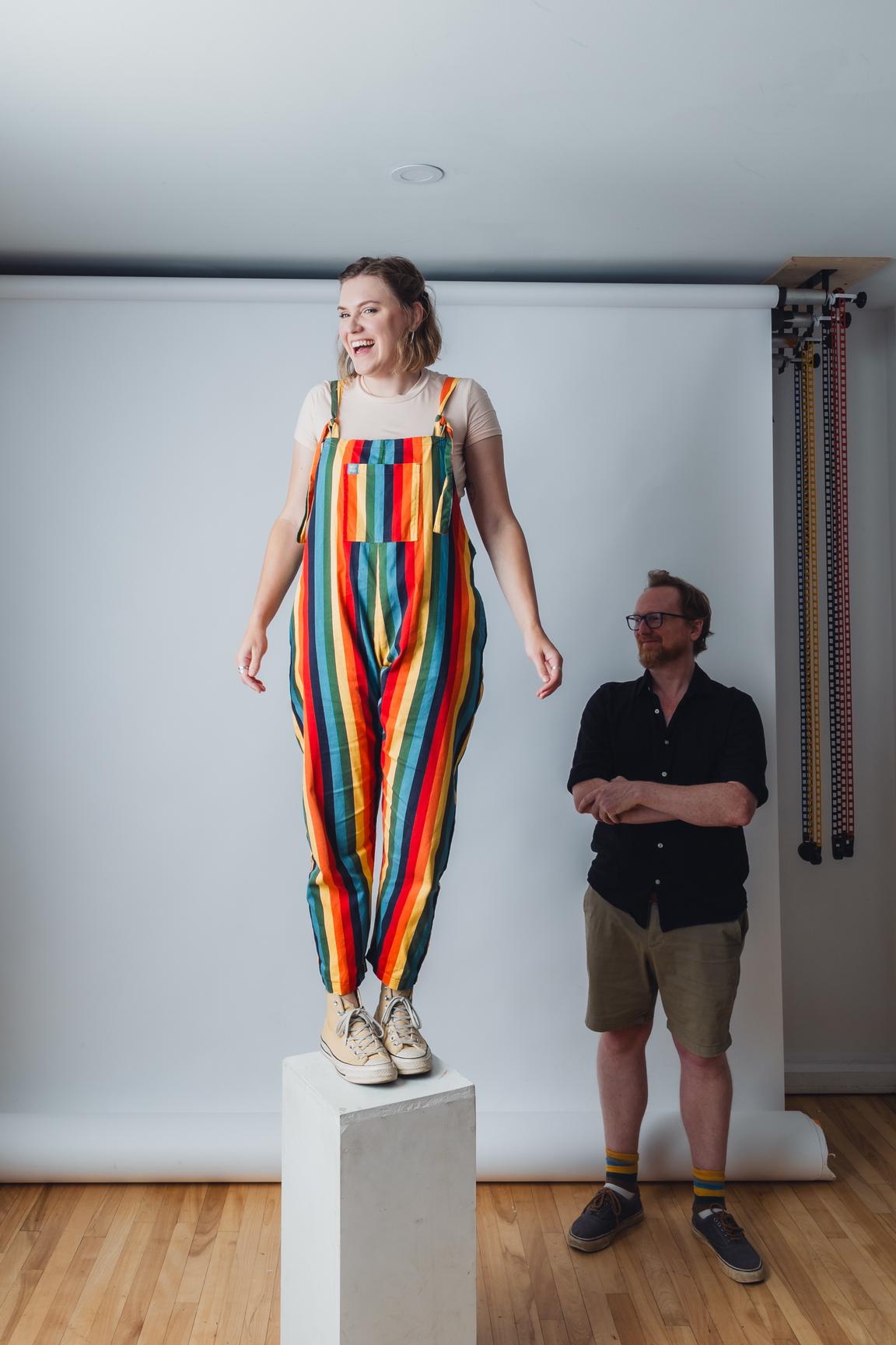 In a photography studio, with hardwood floor. A tall paper photographer's background is unrolled on the wall. Alex stands to the right with his arms crossed, in a shadow. In the centre of the image, Sophie stands on a tall rectangular plinth, in a playful pose.