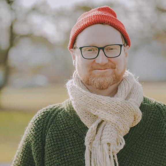 A picture of Alex McLean. He is outside, there are blurry trees and grass behind him. He wears a green sweater, a red toque and has a jaunty scarf tied around his neck. 
