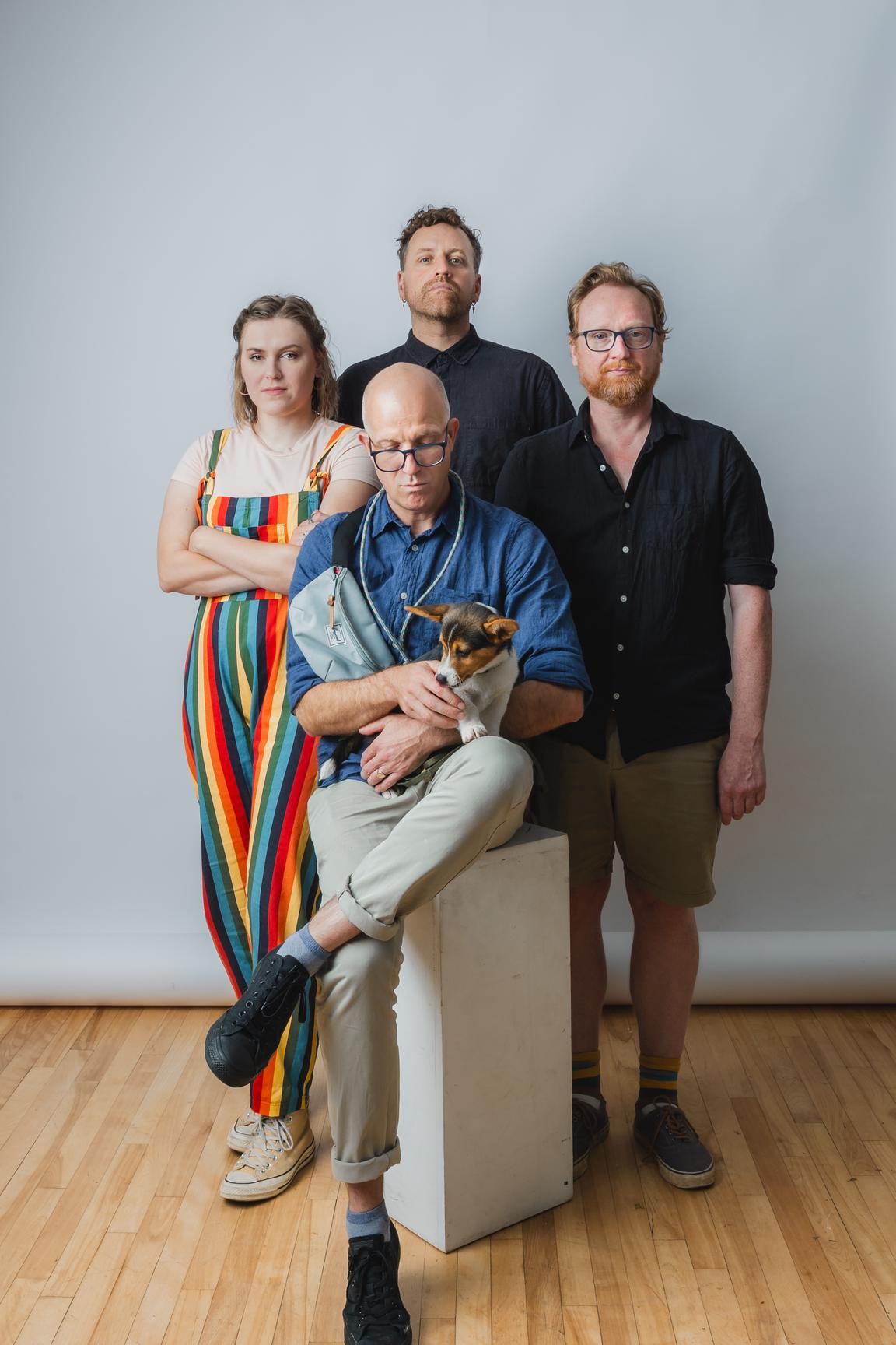 A white/grey photography background. The team stands in a diamond formation, with Ben Stone seated in front on a white plinth. He is holding a corgi puppy named Mabel, and is looking down at her. Clockwise from Ben, to the right, Alex stands with his hands at his sides. Beside him at the top of the diamond, Stewart stands with his head peaking up over Ben's. To the left of the photo, Sophie Stands with her arms crossed. They all look rather serious, but it is a playful pose.