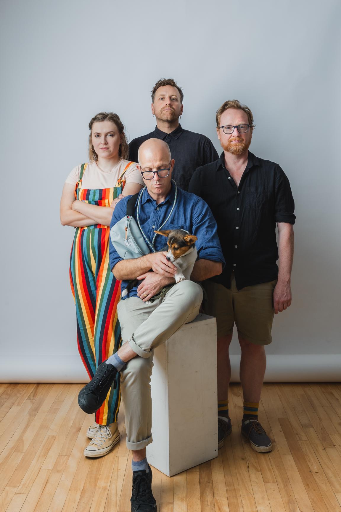 A white/grey photography background. The team stands in a diamond formation, with Ben Stone seated in front on a white plinth. He is holding a corgi puppy named Mabel, and is looking down at her. Clockwise from Ben, to the right, Alex stands with his hands at his sides. Beside him at the top of the diamond, Stewart stands with his head peaking up over Ben's. To the left of the photo, Sophie Stands with her arms crossed. They all look rather serious, but it is a playful pose. 