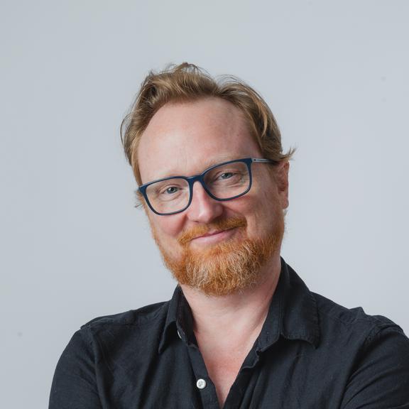 A picture of Alex McLean. He is wearing a blue button-up shirt. He is standing with his arms crossed, his head slightly cocked to one side, and has a pleasant smile on his face. The background is white. 