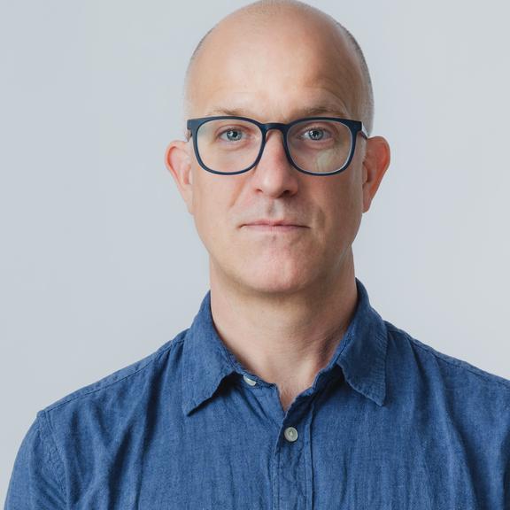 A picture of Ben Stone. From mid torso up. He is wearing a light blue button up shirt. He is squared perfectly to the camera. He has a neutral expression on his face, with the slightest hint of a smile in his eyes.  