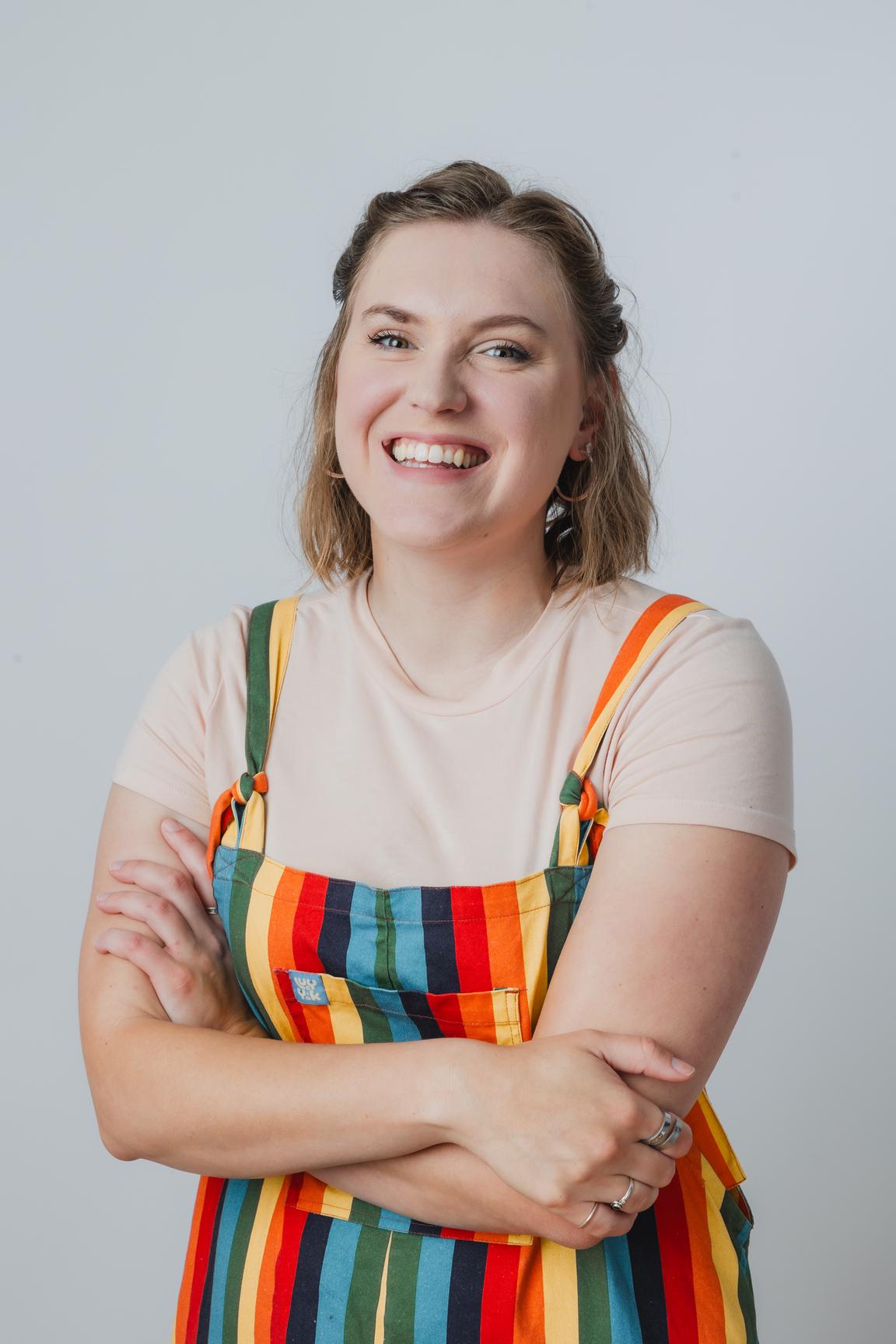 A picture of Sophie Schade. She is standing with her arms crossed. She is wearing a set of rainbow patterned overalls, with a white t-shirt underneath. She appears to have been caught mid-laugh.