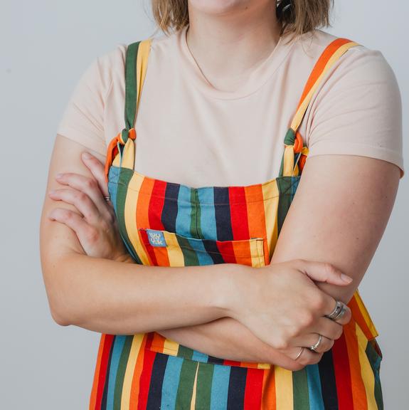 A picture of Sophie Schade. She is standing with her arms crossed. She is wearing a set of rainbow patterned overalls, with a white t-shirt underneath. She appears to have been caught mid-laugh. 