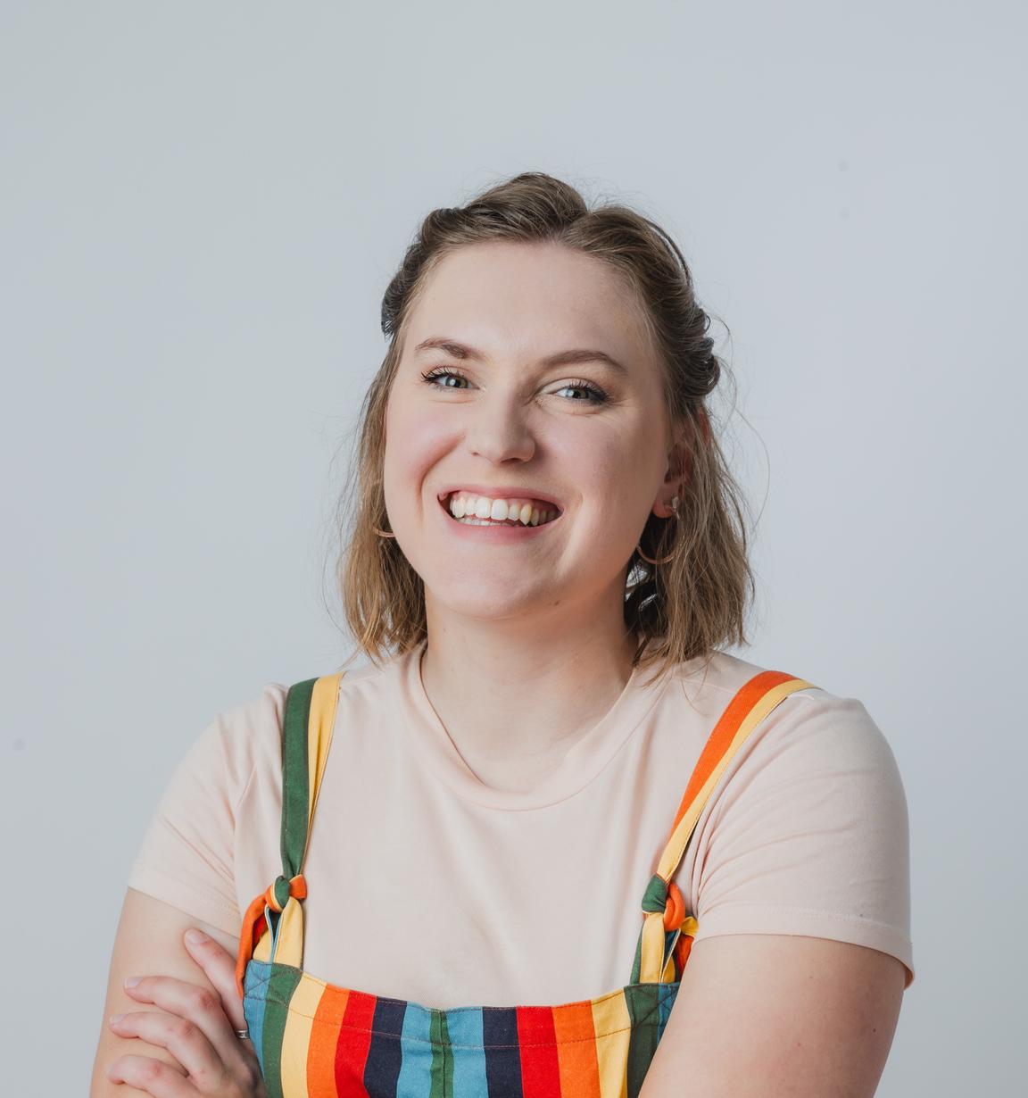 A picture of Sophie Schade. She is standing with her arms crossed. She is wearing a set of rainbow patterned overalls, with a white t-shirt underneath. She appears to have been caught mid-laugh. 