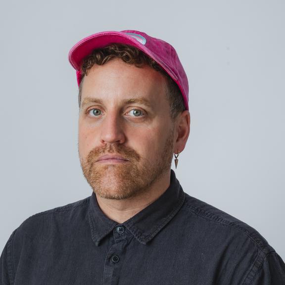 A picture of Stewart Legere. He is slightly angled, looking at the camera. He is wearing a dark blue short-sleeved button up shirt. He is wearing a pink ballcap with a Trans flag on the front. He has short, reddish stubble, and a neutral expression that indicates he doesn't love having his picture taken. 